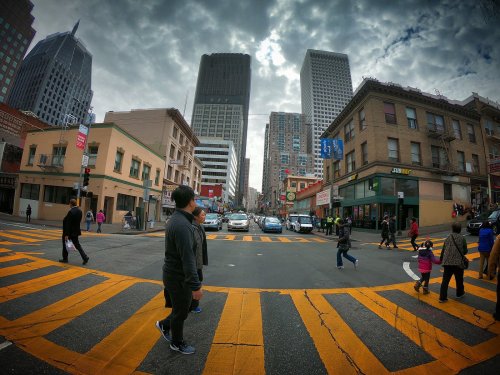 A busy urban intersection with pedestrians crossing, tall buildings, and a cloudy sky in the background.