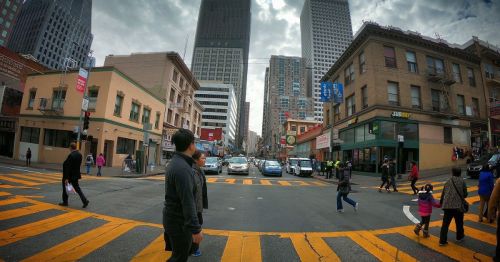 A busy city street with pedestrians crossing, tall buildings, and traffic in the background under a cloudy sky.