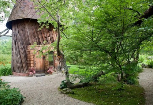 A rustic wooden building surrounded by lush greenery and a gravel path, creating a serene outdoor setting.