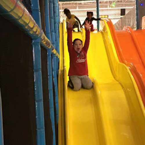 Two children slide down bright yellow slides in a colorful play area, with one child raising their hands in excitement.