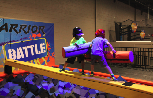 Two kids in helmets engage in a playful battle on a foam pit, using colorful padded poles.