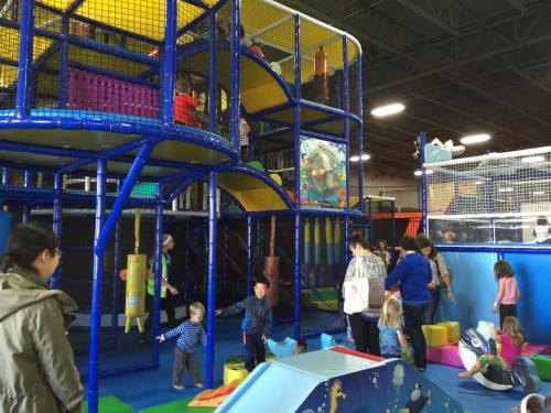 Children play in a colorful indoor playground with climbing structures and soft play areas, while adults supervise nearby.