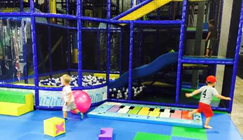 Two children play in a colorful indoor playground with a ball, slide, and soft play structures.