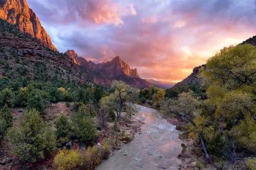 A scenic view of a river winding through a canyon, surrounded by trees and mountains under a colorful sunset sky.