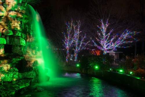 A waterfall illuminated in green, surrounded by trees adorned with colorful lights at night.