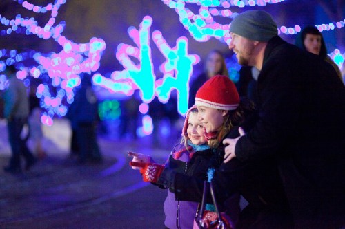 A man and a girl in winter attire admire colorful holiday lights in a festive outdoor setting.