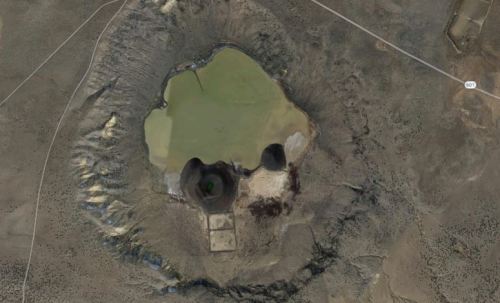 Aerial view of a large, circular crater with a greenish pond at its center, surrounded by rocky terrain.