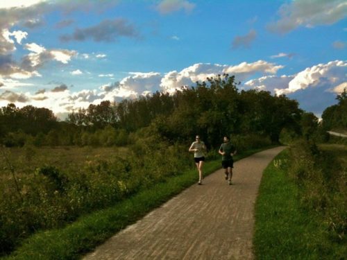 Two runners jog along a scenic path surrounded by greenery and a blue sky with fluffy clouds.