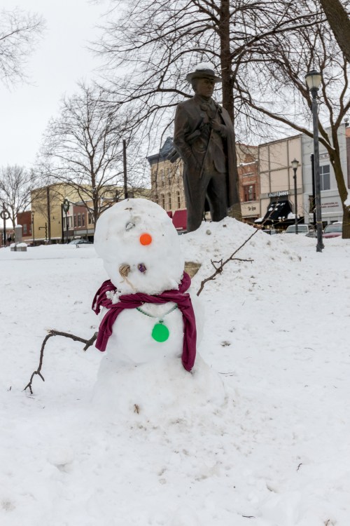 A snowman with colorful buttons and a scarf stands in a snowy park, with a statue and buildings in the background.