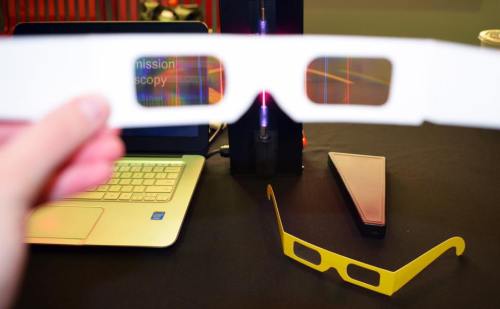 A hand holds white 3D glasses in front of a laptop and a yellow pair of glasses on a table.