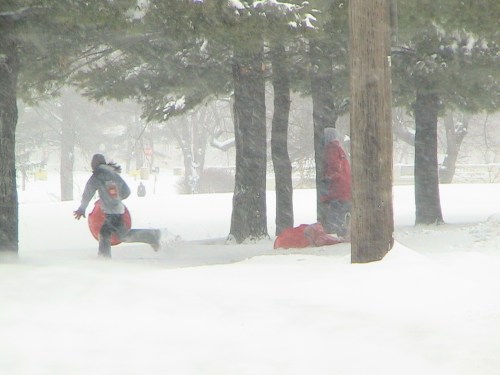 A person runs through heavy snow, holding a sled, while another person stands nearby among snowy trees.