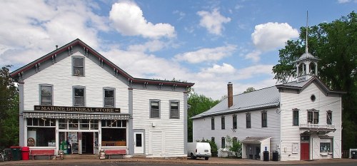 Historic white buildings with a general store and a small chapel, set against a blue sky with fluffy clouds.