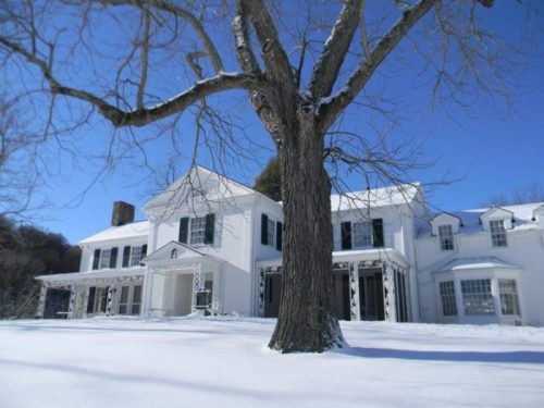 A large white house surrounded by snow, with a bare tree in the foreground and a clear blue sky above.