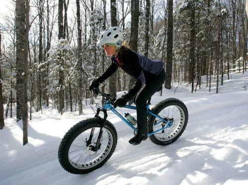 A person rides a fat bike through a snowy forest, wearing a helmet and winter gear. Snow-covered trees surround them.