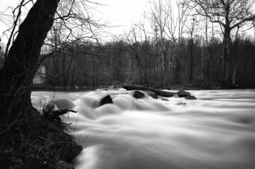A black and white image of a flowing river with smooth water, surrounded by trees and rocky banks.