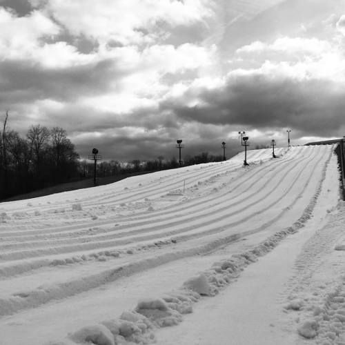 A snowy hill with ski tracks under a cloudy sky, surrounded by bare trees.