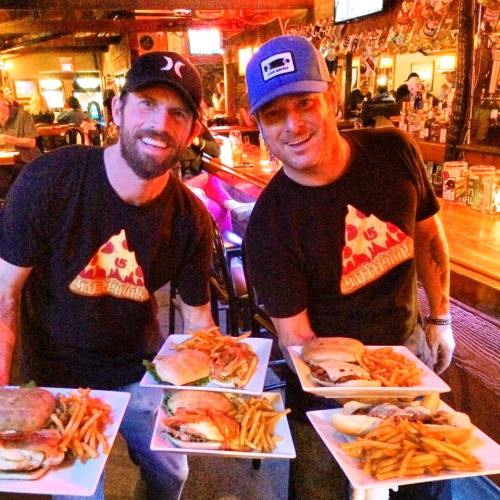 Two men in matching pizza-themed shirts hold plates of burgers and fries in a lively bar setting.