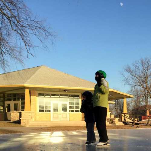 A child and an adult stand on a frozen surface near a building, with a clear blue sky and a crescent moon in the background.