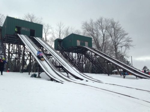 Two snow-covered sledding hills with green structures at the top, surrounded by trees under a cloudy sky.