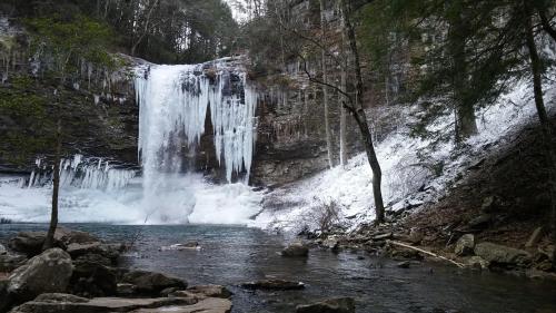 A frozen waterfall surrounded by icy rocks and trees, with a calm river flowing in the foreground.
