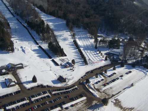 Aerial view of a snowy ski resort with slopes, parking lot, and surrounding trees.