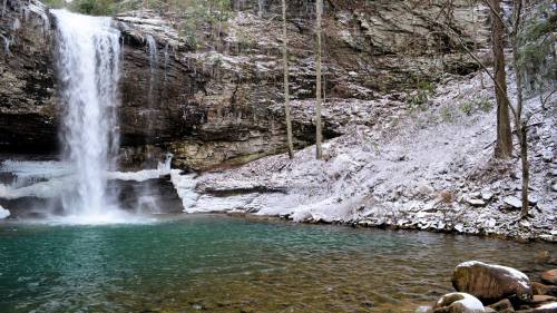 A serene waterfall cascades into a turquoise pool, surrounded by snow-dusted rocks and trees in a tranquil forest setting.