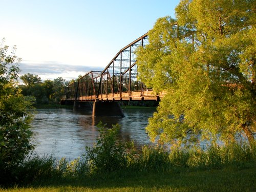 A historic metal bridge spans a river, surrounded by lush greenery and a clear blue sky.