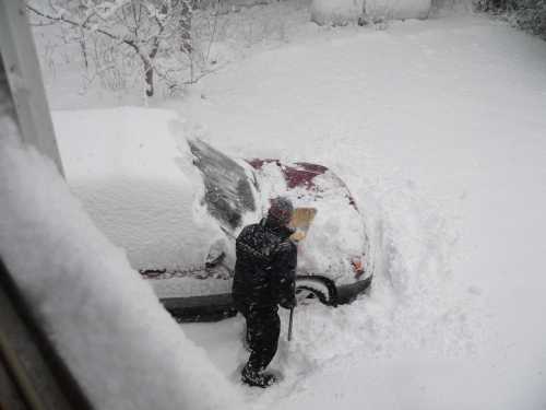 A person shovels snow off a car buried in a heavy snowfall, with a snowy landscape in the background.