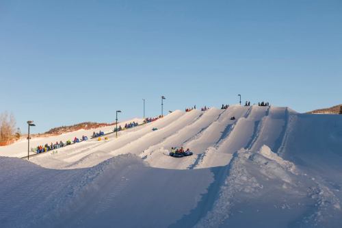 A snowy hill with people sledding down and others waiting in line at the top, under a clear blue sky.