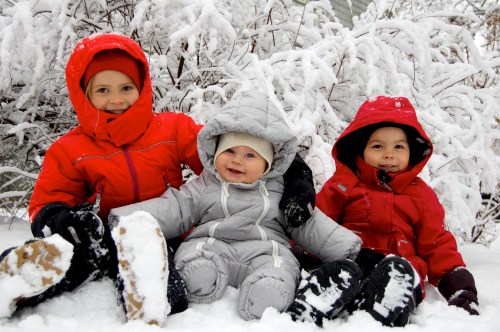 Three children in colorful winter coats sit in the snow, smiling amidst a snowy landscape with frosty branches.