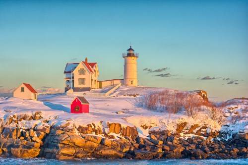 A snowy landscape featuring a lighthouse and a red shed on rocky terrain, illuminated by warm sunset light.