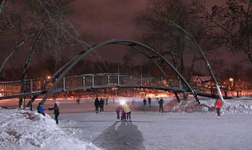 A snowy ice rink at night with skaters under a bridge, surrounded by trees and city lights in the background.