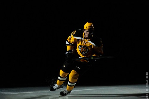 A hockey player in a yellow and black jersey skates on the ice, illuminated against a dark background.
