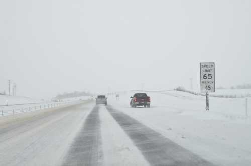 A snowy road with low visibility, featuring two vehicles and a speed limit sign indicating 65 mph, minimum 45 mph.