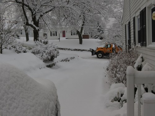 A snow-covered driveway with a snowplow clearing the path, surrounded by trees and houses in a winter scene.