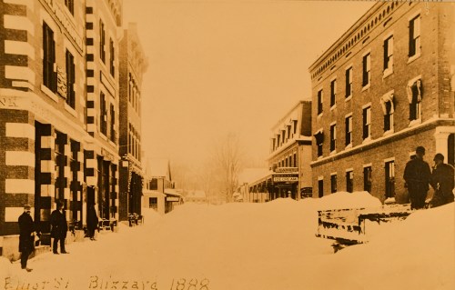 Historic photo of a snowy street scene from the 1888 blizzard, featuring buildings and people amidst deep snow.