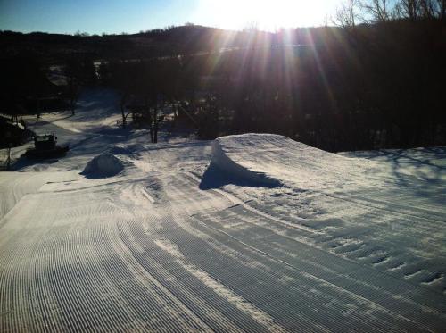 A snowy landscape with groomed ski slopes and jumps, illuminated by sunlight in the background.