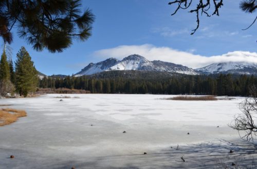 A frozen lake surrounded by pine trees and snow-capped mountains under a clear blue sky.