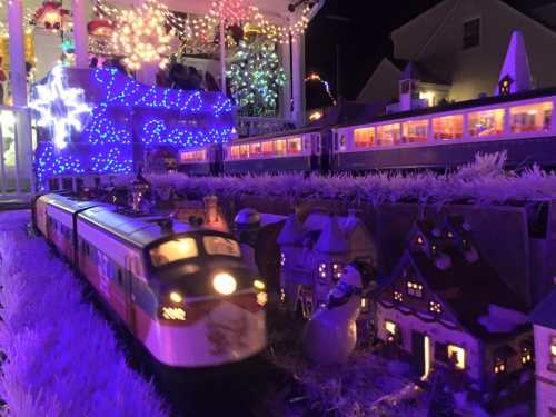 A festive train set with colorful lights, passing by miniature houses and a snowy landscape during the night.