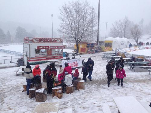 A snowy outdoor scene with people gathered around wooden stumps, food trucks in the background, and trees nearby.