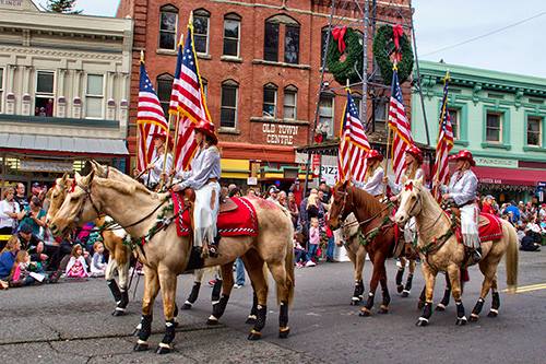 A parade featuring riders on horses adorned with American flags, passing by a crowd and colorful storefronts.