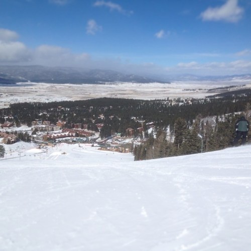 A snowy mountain landscape with a view of a ski resort and surrounding trees under a blue sky.
