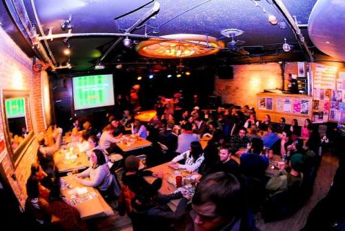 A lively bar scene with a crowded audience, tables filled with people, and a stage in the background.
