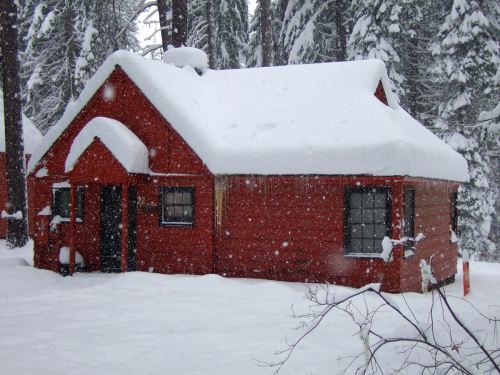 A red cabin covered in snow, surrounded by tall trees in a winter landscape. Snowflakes are falling gently.