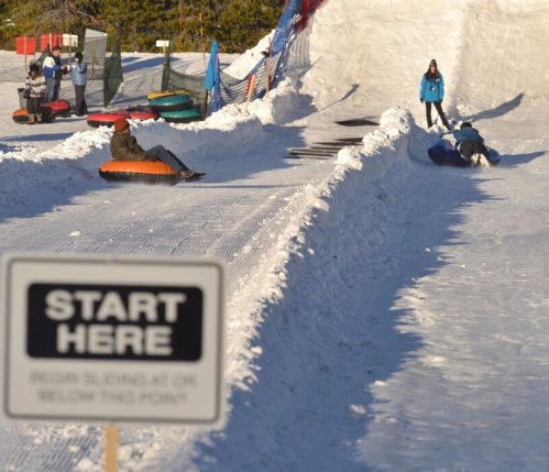 A snowy tubing hill with people sliding down and a sign that says "START HERE" in the foreground.