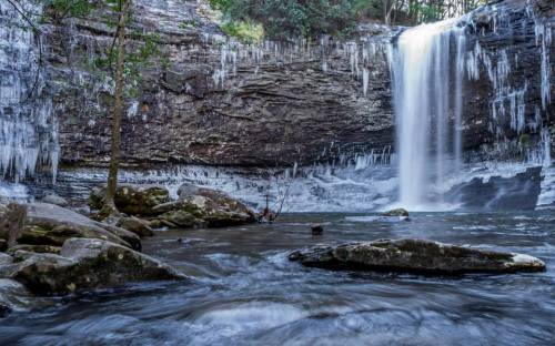 A serene waterfall cascades over rocky cliffs, surrounded by icicles and a flowing river.
