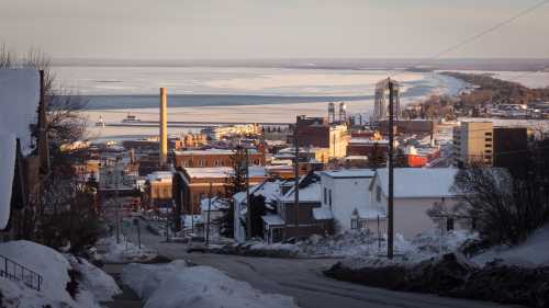 A snowy hillside view of a town by the water, with buildings and a frozen lake in the background under a clear sky.