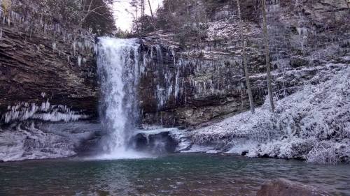 A frozen waterfall cascades into a serene pool, surrounded by icy rocks and snow-covered trees.