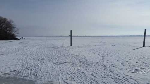 A snowy landscape with a frozen lake, featuring two poles in the foreground and a cloudy sky above.