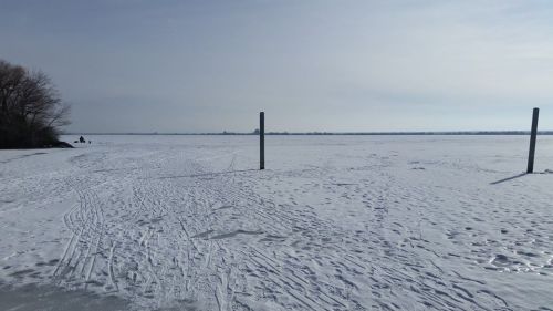 A snowy landscape with a frozen lake, featuring two poles in the foreground and a cloudy sky above.